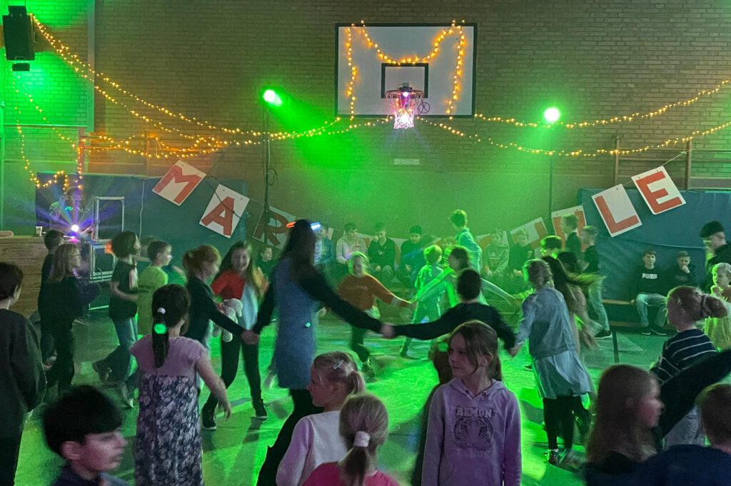 Zur Belohnung ab zur Party Bereits zum vierten Mal in Folge organisierte die Schulpflegschaft der Marienschule Hauenhorst am Tag der Ausgabe der Halbjahreszeugnisse zur Freude aller die beliebte „Zeugnisdisco“. Ob Erst- oder Viertklässler, Dancing-Queen oder eher Tanzmuffel, ob gutes oder weniger gutes Zeugnis – alle waren da. Und natürlich hat sich auch in diesem Jahr das Kommen mal wieder mehr als gelohnt. Die vom erfolgreichen Crowdfunding angeschaffte Soundanlage und die tanzwütigen Kids brachten die Halle zum Beben. DJ Sven spielte was die Kinder sich wünschten. Discolichter wohin man schaute, eine gut funktionierende Nebelmaschine und der Lieblingsflashmob der Marienschülerinnen und Marienschüler unter der Leitung der momentan abgeordneten Kollegin Maike Hüls sorgten für allerbeste Stimmung. Zwischendurch konnten sich alle an ihren vorbereiteten Tischen mit Getränken und Snacks stärken. Für die Ausgabe der Hotdogs ging man Jahrgangsweise zum discomäßig verkleideten Team der Schulpflegschaft. Auch hier sorgte eine perfekte Organisation für einen reibungslosen Ablauf. Zwei Stunden waren schnell vorbei, nachwirken wird die Disco jedoch auf jeden Fall noch lange. Und: Erfolg verpflichtet. Ganz bestimmt dürfen sich die Kids in Hauenhorst auch im Schuljahr 2026/27 am Tag der Halbjahreszeugnisse auf die fünfte Auflage der „Marienschulzeugnisdisco 2027“ freuen.