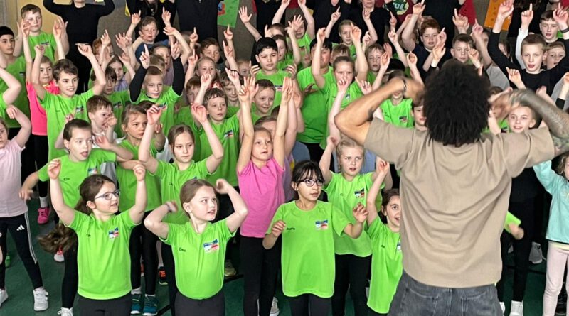 Bewegung, die Spaß macht „Liebes Team der Marienschule, glauben Sie uns, wir erreichen mindestens 95% ihrer Kinder und es werden alle ganz viel Spaß haben, ganz bestimmt.“ „So ähnlich könnte man die Antwort auf unsere erste Anfrage bei www.wirbewegenschule.de“ wiedergeben“, so das Schulleitungsteam der Marienschule Hauenhorst, Alexandra Nieweler und Christian Hülsmann. Anlässlich der Auszeichnung als „BuG-Schule“ (Bildung und Gesundheit“) suchte man an der Marienschule nach einem Projekt, welches perfekt zu den Hauptaspekten des NRW-Landesprojektes „Bildung und Gesundheit“ passt. „Wir bewegen Schule“ passt tatsächlich wie der Deckel auf den Topf. Viel besser hätten wir uns nicht entscheiden können, denn wesentliche Inhalte von BuG wurden in dieser Projektwoche zu 100% getroffen,“ so Alex Nieweler, die von diesem Projekt gehört hatte und es unbedingt nach Hauenhorst an die Marienschule holen wollte. „Und als dann die Zertifizierung anstand, und alles so stimmig schien, war auch unser „Chef“ begeistert und los ging es“, schmunzelt Nieweler noch heute von den Anfängen der Idee vor ca. 8 Monaten. Das Bildungsportal NRW schreibt übrigens folgendes über das Landesprojekt „Bildung und Gesundheit“: „Die Schule sollte sowohl für Lehrkräfte als auch für Kinder und Jugendliche ein lebenswerter Arbeitsplatz sein. Dies gelingt am besten, wenn sich alle an der Entwicklung ihrer Schule zur „guten gesunden Schule“ beteiligen. Bei der Entwicklung des eigenen Schulprogramms spielt das Thema Gesundheitsförderung für Kinder und Jugendliche sowie Lehrkräfte eine entscheidende Rolle. Gesundheitsförderung bezieht alle Bereiche des schulischen Lebens ein und wird als schulischer Entwicklungsprozess auf allen Ebenen der Bildungsverwaltung, etwa durch Fachberater und Dezernentinnen und Dezernenten für Gesundheit bei den Bezirksregierungen mit dem Ziel der Verbesserung der Bildungsqualität durch eine gesteigerte Gesundheitsqualität unterstützt. Gute Gesundheit wirkt sich in der Schule in vielerlei Hinsicht positiv aus: Herausforderungen werden besser bewältigt, das Konzentrieren fällt leichter und die Leistungsfähigkeit steigt. Schulen können ihr Engagement zur Weiterentwicklung eines gesundheitsfördernden Schulklimas durch Teilnahme am Landesprogramm Bildung und Gesundheit zertifizieren lassen und weiter ausbauen.“ Genau das hat die Marienschule in den vergangenen zwei Jahren fleißig gemacht und am Freitag war es dann soweit. Das neue „Leitbild“ der Marienschule wurde allen anwesenden Gästen (und das waren bei zwei ausverkauften Vorstellungen eine ganze Menge) durch die Kids der Schule vorgestellt und Ingrid Deupmann, Regionalkoordinatorin bei BuG war mit dem Zertifikat in Form eines Hausschildes zu Besuch und zum Gratulieren persönlich vorbeigekommen. Sehen konnten sie und auch die vielen Gäste zwei bewegende und mitreißende Vorführungen individueller Klasseninszenierungen bei cooler Musik, ganz viel Tanz, starkem Ausdruck, anspruchsvoller Akrobatik und mitreißendem Rhythmus.“ An den Tagen zuvor hatten alle acht Klassen täglich 90 Minuten Tanzunterricht bei Amadin, Immanuell und Niclas vom Team „Wir bewegen Schule“ gehabt und beeindruckende Darbietungen und Choreografien erlernt. Was bei den Generalproben am Freitagmorgen noch den letzten Schliff bekommen hatte, klappte am Nachmittag nahezu tadellos. Begeisterte Eltern, Großeltern und Freunde staunten nicht schlecht, sparten nicht mit Applaus und zollten sowohl den drei Tänzern als auch dem Kollegium der Marienschule, welches zur großen Freude aller ebenfalls einen Tanz eingeübt und zu Beginn der Shows aufgeführt hatte, großen Tribut. Alle Kinder erlebten in dieser Woche, wie sich ein respektvoller und wertschätzender Umgang auf die Klassengemeinschaft, die eigene Motivation und die gemeinsame Begeisterung auswirkt. Die Kinder unterstützten und bejubelten sich schon im Training. Jedes Kind fühlte sich gesehen, durfte sich ausprobieren und konnte sein individuelles Können einbringen. Die Woche „war nicht nur ein voller Erfolg, sondern ein Erlebnis, das unsere Schulgemeinschaft bereicherte“, heißt es aus dem Kollegium, welches voll des Lobes war.