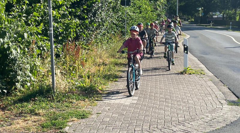 Wandertag zur Kreyenburg – ja, mir san mit dem Radel do Die Woche vor den Sommerferien beinhaltet fast immer und überall in Schulen einen klassischen Wandertag. Aber was heißt schon klassisch? Nur noch selten wird tatsächlich gewandert. An der Marienschule in Hauenhorst herrscht hier rege Abwechslung. In dem einen Jahr geht es zu Fuß zur Hasenhöhle und mit dem Bus zurück, in einem anderen Jahr wird mit Germania Hauenhorst ein großes Fußballturnier auf die Beine gestellt und dann gibt es auch noch den Wandertag zum Spielplatz Kreyenburg. Der stand in diesem Jahr mal wieder auf dem Plan. Da kam der Schulleitung die Idee, diesen Wandertag im Rahmen des Stadtradelns mal so richtig zum Kilometersammeln zu nutzen. Gute Idee, fand auch die Schulpflegschaft und war sofort mit im Boot. Aber: Mit allen Klassen??? Nicht in der Woche vor den Sommerferien??? Und wie soll das überhaupt funktionieren. Das OK war gegeben und Probleme sind zum Lösen da, also entschied sich das Schulleitungsteam für die Durchführung. Das Kollegium war begeistert, verschiedenste Ideen wurden gesammelt, die Polizei mit ins Boot geholt und alle Eltern zum Üben des Radfahrens aufgerufen. Und lange Rede, kurzer Sinn: Am Dienstag, also nicht im Rahmen des Stadtradelns, war es soweit. Bei bestem Wetter radelten im Schnitt pro Klasse 20 Kinder und 5 Erwachsene zeitlich versetzt von der Marienschule zur Kreyenburg und im Mittag dann auch wieder zurück. Verkehrssicherheitsberater Andreas Kröger instruierte zu Beginn der Fahrten an der Schule alle Teilnehmer und führte jede der acht Gruppen sicher durch Hauenhorst bis zum „entspannten“ Hessenweg. Ab hier hatten die beiden „Dorfpolizisten“ für Neuenkirchen und Hauenhorst/Catenhorn, Frank Heymann und Udo Wilmer, die beste Strecke ausgearbeitet und für alle mit Wegpfeilen präpariert. So kamen die Gruppen ganz entspannt und nahezu verkehrsfrei nach Neuenkirchen, wo die beiden Bezirkspolizisten die Klassen dann noch nacheinander sicher durch die einzige Engstelle leiteten. Besser geht es nicht. An der Kreyenburg angekommen wurden Autoparkplätze zu Fahrradabstellplätzen, schließlich mussten über 200 Räder abgestellt werden, und dann begann ein buntes Treiben auf diesem tollen Spielplatz. Auch hier fand die Schule, dank des tollen Einsatzes zweier Kolleginnen aus Neuenkirchen, ideale Bedingungen vor. Und als dann um 10:30 Uhr auch noch der Eiswagen vorfuhr, war allen klar: Das ist nicht mehr zu toppen. Auch der Rückweg lief dann mit lediglich einer Fahrradpanne und einem kleineren Sturz ähnlich unkompliziert wie der Hinwege. Die Schulleitung der Marienschule zeigte sich am Ende des Tages sehr erleichtert und war voll des Lobes für dieses Projekt: „Unser Mut wurde belohnt. Ob Erstklässler oder Viertklässler mit bestandener Fahrradprüfung – heute haben alle aktiven Radler gezeigt, das solch ein ambitioniertes Vorhaben gelingen kann. Die Kids waren überragend, die vielen helfenden Eltern der Hammer und die Unterstützung durch die Polizei vorbildlich“, so Christian Hülsmann und Alex Nieweler, das Stadtradel-Captain-Team der Marienschule, die solch eine Kilometerleistung (immerhin gut 2500km an einem Tag) beim nächsten Mal doch gerne während des Stadtradelns realisieren möchten. „Schauen wir mal, was uns 2027 dazu einfällt“, so die beiden mit einem verschmitzten Lächeln im Gesicht.